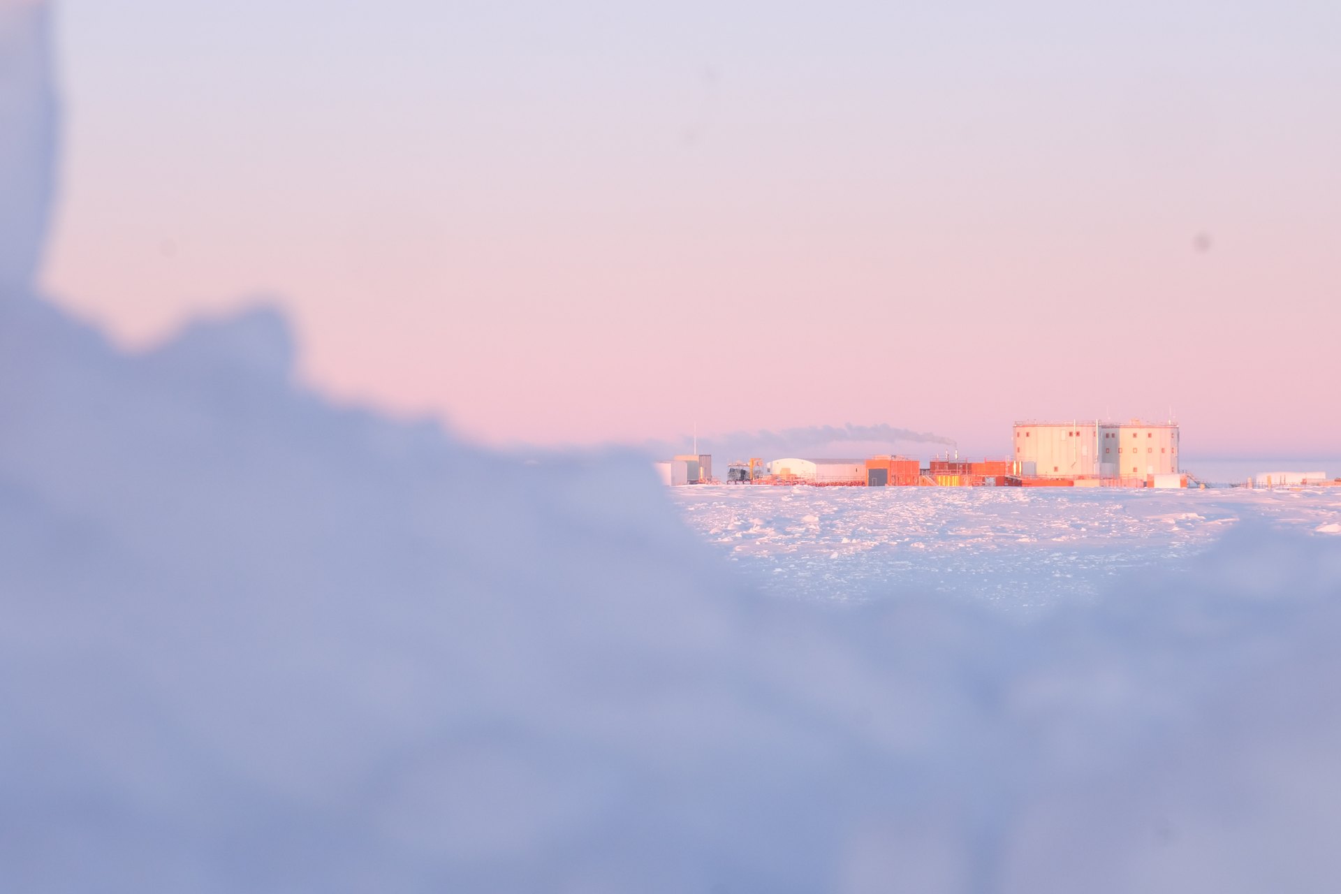 La estación Concordia, dos edificios cilíndricos y varios contenedores naranjas, vista de lejos y a ras de suelo sobre la llanura nevada del Dome C antártico, con el cielo rosado del sol bajo y una loma de nieve desenfocada en primer plano.
