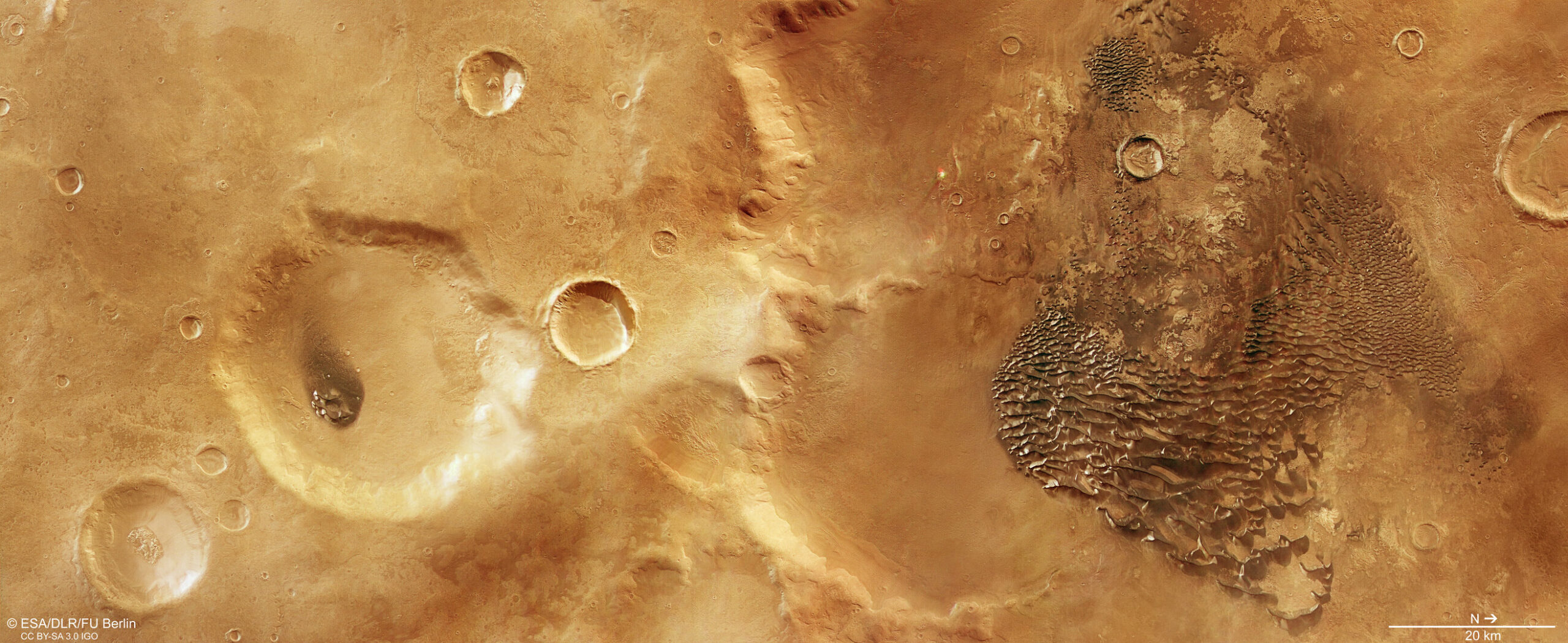 Vista cenital de una llanura de Marte en tonos naranjas y ocres, sembrada de cráteres de impacto de todos los tamaños. Una cresta clara cruza la escena de arriba abajo por el centro y, a la derecha, un campo de dunas oscuras y apretadas forma un dibujo de olas con manchitas blancas encima.