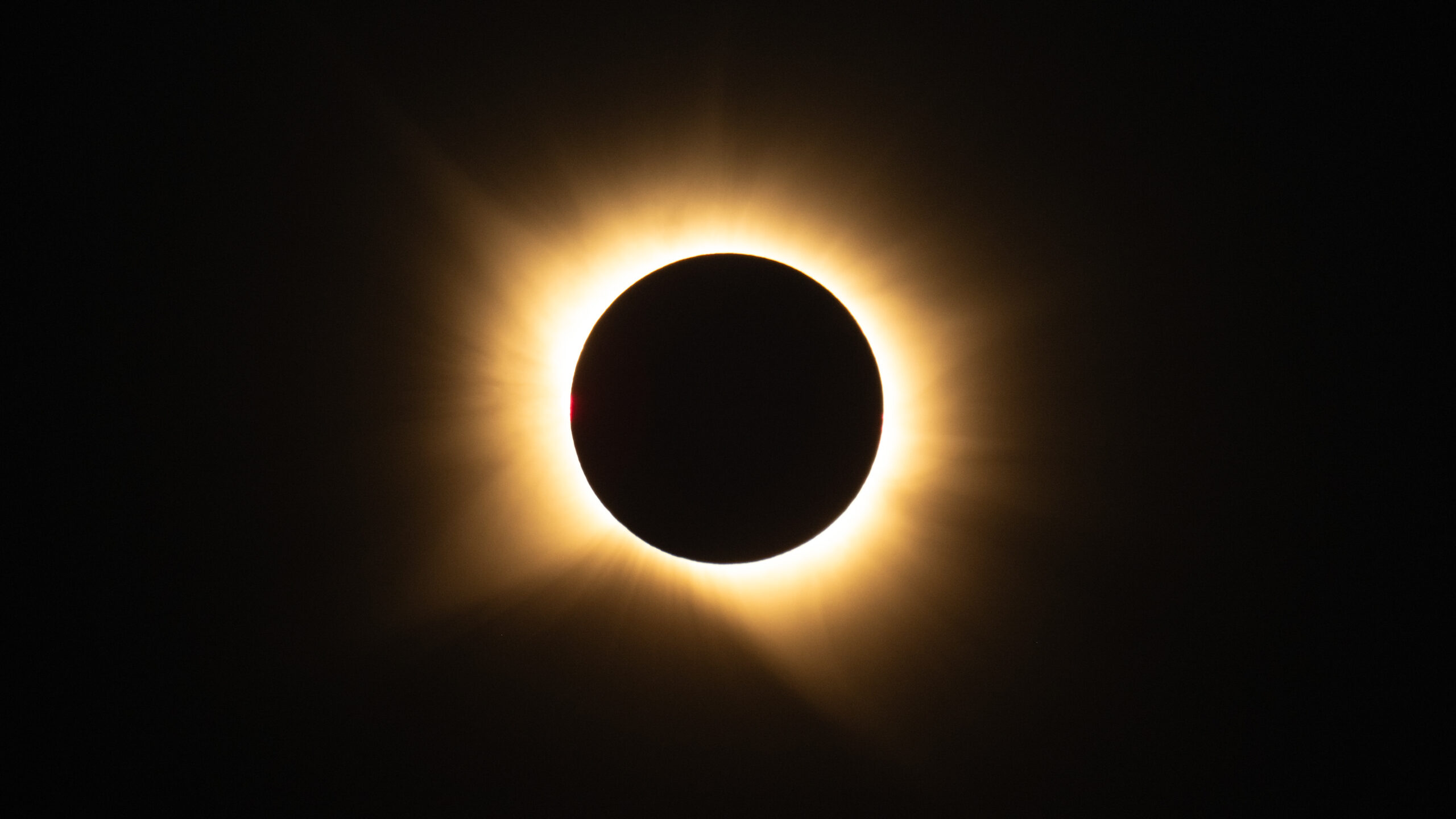 El disco negro de la Luna tapa por completo al Sol y deja alrededor un anillo de luz amarilla y anaranjada, la corona solar, sobre un cielo oscuro. En el borde izquierdo del disco asoma un punto rojo.