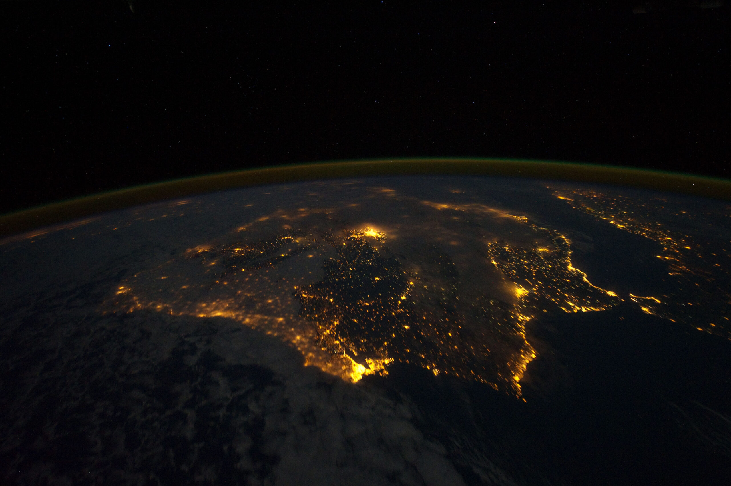 La Península Ibérica de noche vista desde la órbita terrestre. Sobre el fondo negro del planeta, las luces de las ciudades dibujan en dorado anaranjado el contorno de toda la costa y una malla densa de puntos por el interior. Cerca del centro destaca una mancha compacta y mucho más brillante que el resto, que es Madrid. A la izquierda, un manto de nubes tenuemente iluminadas por la Luna cubre el Atlántico. Arriba, la curva del horizonte terrestre está subrayada por una franja de luminiscencia atmosférica que va del dorado al verde, y por encima solo hay espacio negro con estrellas.