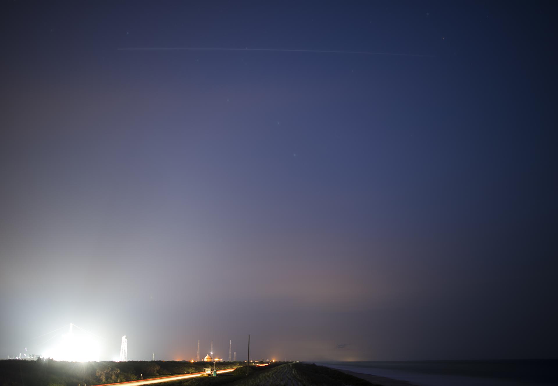 Fotografía nocturna de larga exposición: una raya de luz continua y sin interrupciones cruza el cielo estrellado sobre la silueta iluminada de una torre de lanzamiento. La raya es el rastro que deja la Estación Espacial Internacional durante el minuto que duró la exposición.