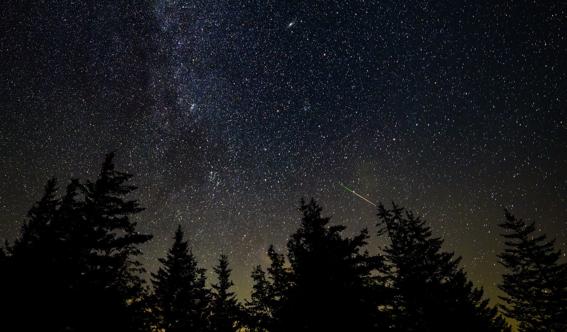 Un meteoro cruza el cielo nocturno como una raya de luz brillante sobre un campo de estrellas, con la banda de la Vía Láctea al fondo y la silueta negra de la línea de árboles en el horizonte.