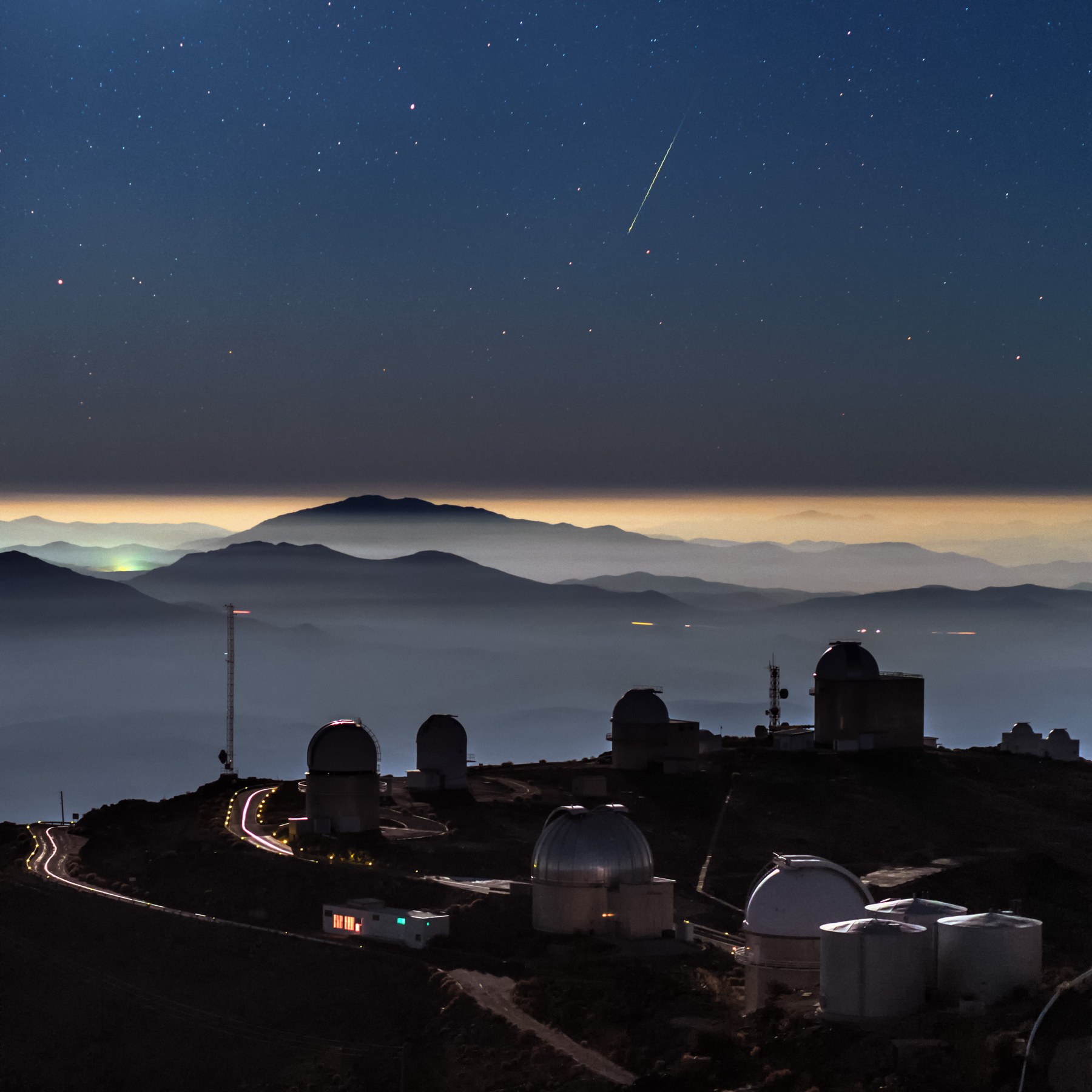 Un meteoro brillante deja una raya de luz verdosa en un cielo estrellado, sobre las cúpulas blancas de un observatorio encaramado en una loma, con un mar de nubes y varias filas de montañas recortadas contra el resplandor del horizonte.