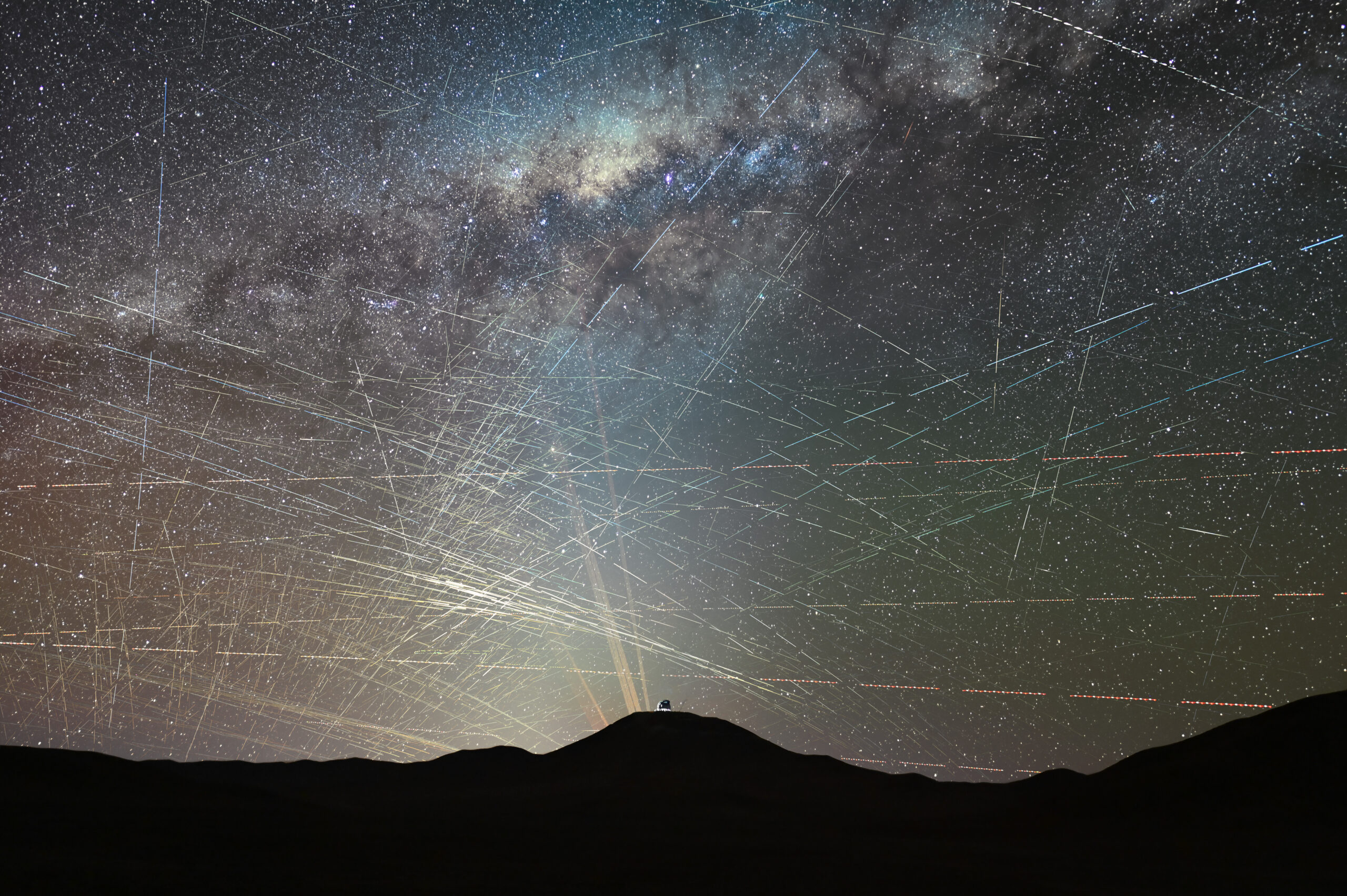 Decenas de estelas rectas de satélites cruzan el cielo estrellado del desierto de Atacama sobre la cúpula del telescopio ELT, todavía en construcción; al fondo, los láseres del observatorio Paranal apuntan al cielo.