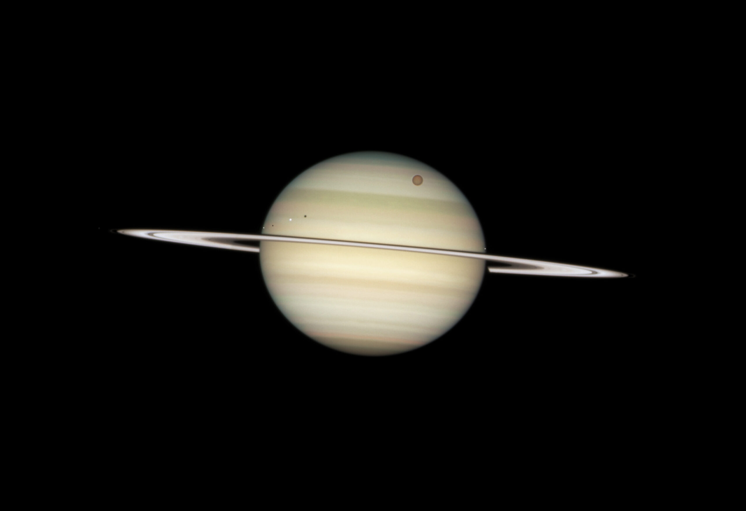 Saturno visto por el Hubble con los anillos casi de canto, reducidos a una línea fina y luminosa que cruza el disco de lado a lado; sobre las nubes del planeta se recortan cuatro de sus lunas, la mayor anaranjada, con sus sombras al lado.