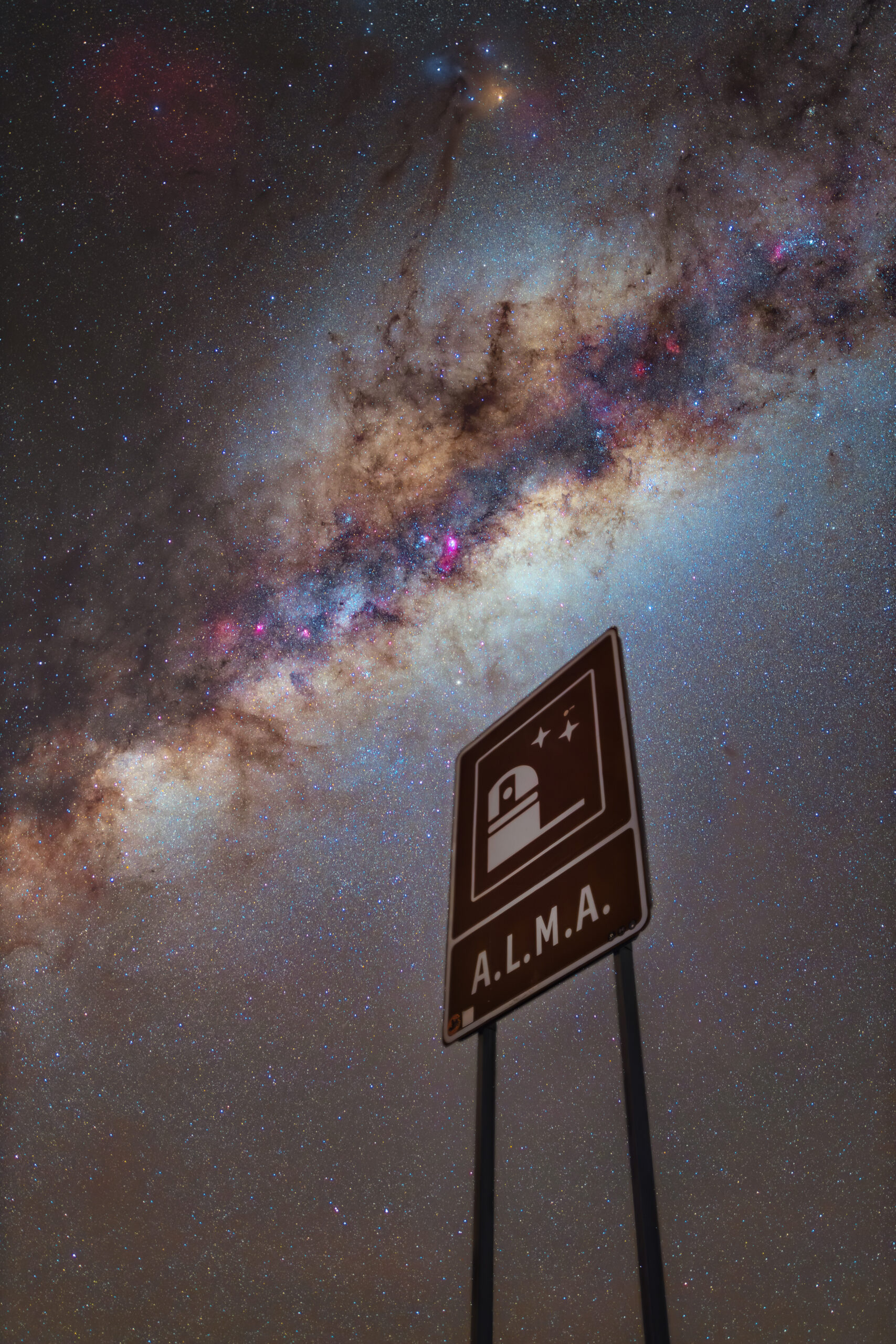 La banda de la Vía Láctea se levanta sobre el desierto de Atacama, de noche y llena de estrellas, detrás de la señal de tráfico que marca la entrada al observatorio ALMA junto a una carretera vacía.