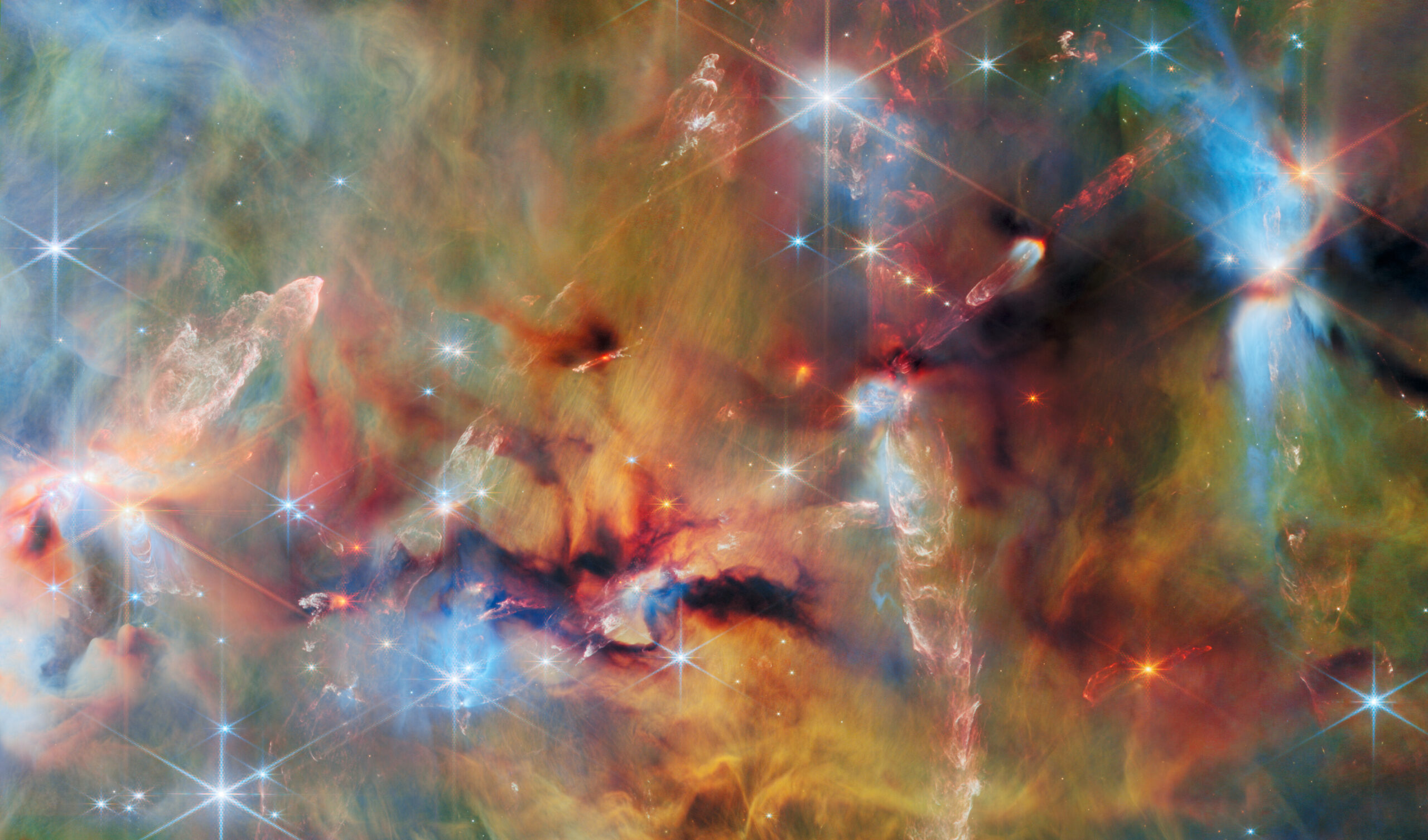 Nube de gas y polvo fotografiada en infrarrojo: capas verdes, amarillas y anaranjadas atravesadas por manchas de polvo oscuro casi negro, decenas de estrellas brillantes de seis puntas y varios chorros rojizos de gas que salen de estrellas en formación.