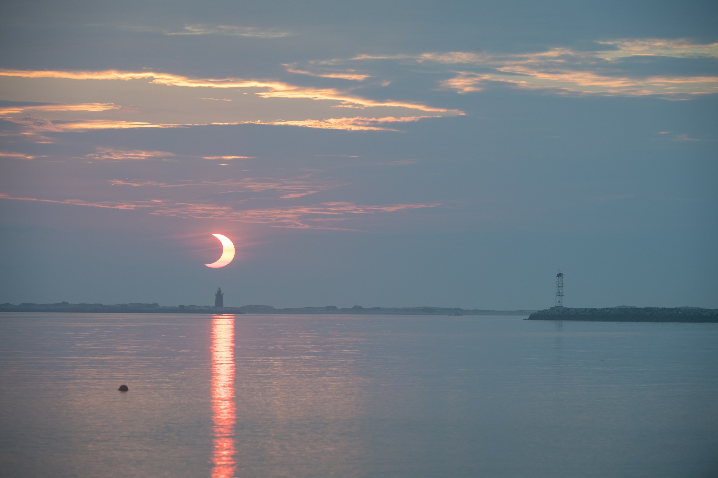 El Sol asoma sobre el horizonte marino con un mordisco oscuro en su borde superior, detrás de la silueta de un faro.