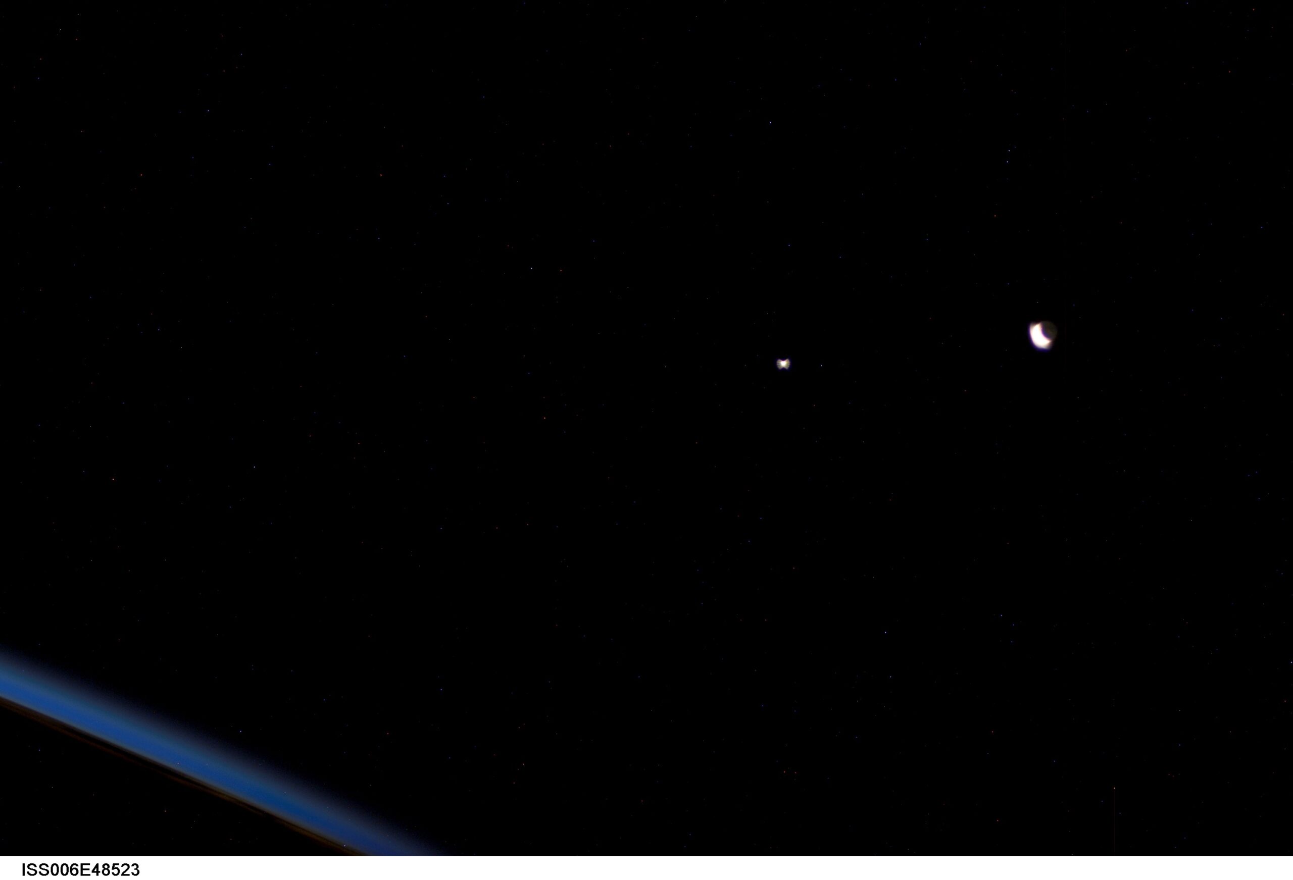 Sobre un cielo negro sembrado de estrellas débiles, la Luna en creciente a la derecha, tan sobrexpuesta que se distingue también su parte oscura, y Venus como un punto blanco brillante en el centro; abajo a la izquierda asoma el arco azul del horizonte de la Tierra visto desde la órbita.