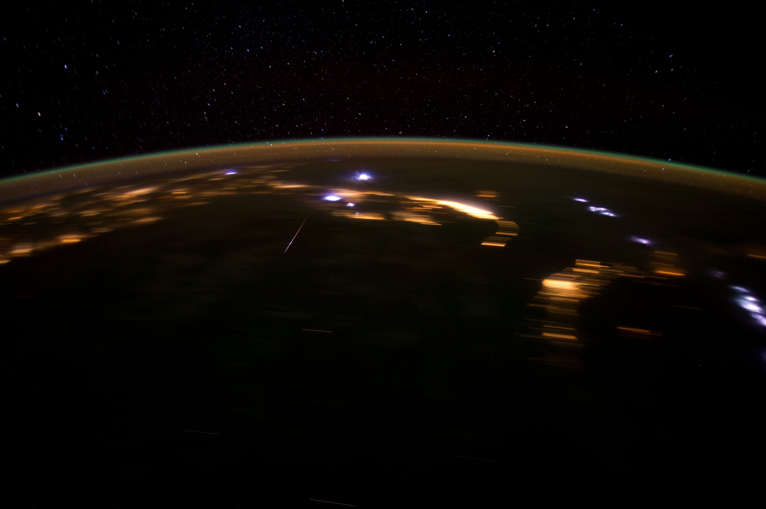 La Tierra de noche vista desde la órbita, con la curva del horizonte y una fina capa de atmósfera anaranjada bajo un cielo lleno de estrellas. Sobre el planeta oscuro brillan las luces anaranjadas de varias ciudades costeras y unos destellos azulados de relámpagos entre las nubes. A la izquierda del centro, un meteoro deja una raya blanca, corta y recta.