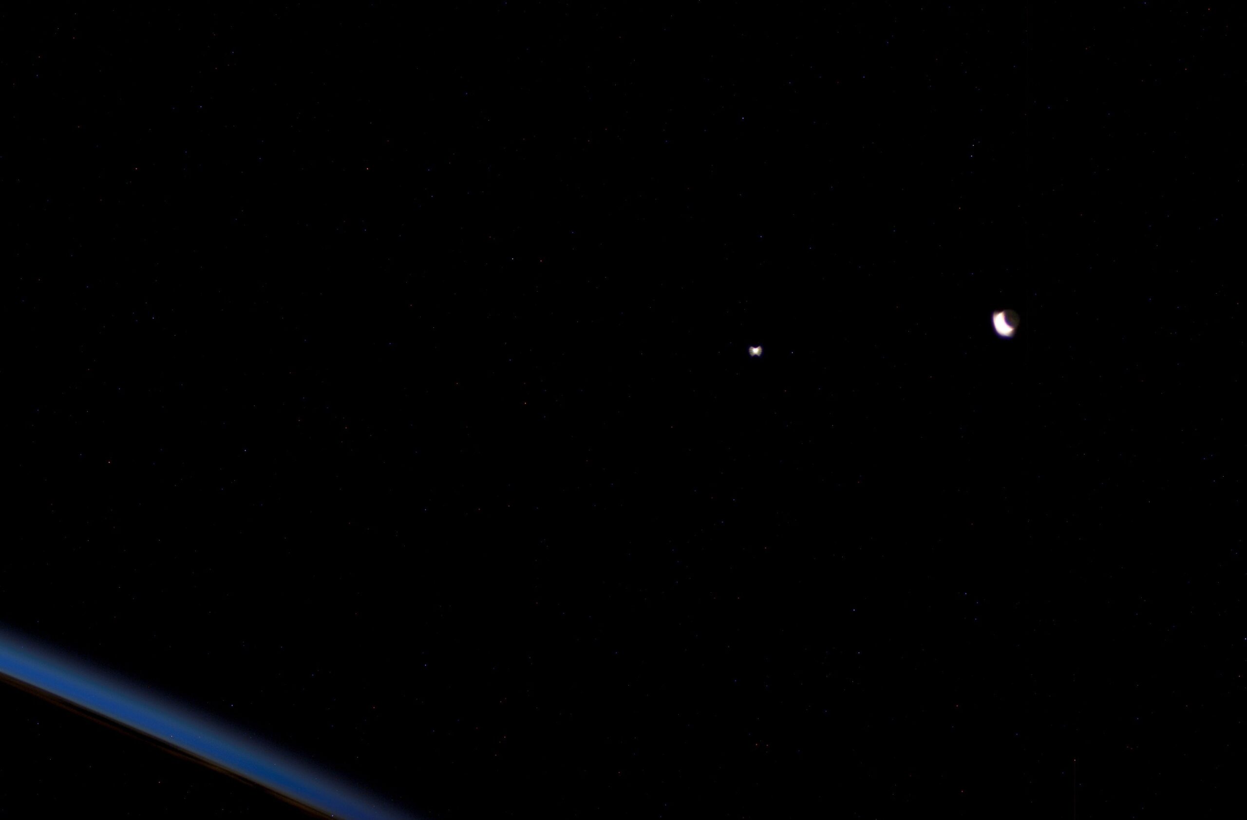 Fotografía casi toda negra: en el centro, un punto blanco muy brillante; a su derecha, una Luna en fase de hoz con la parte oscura del disco apenas visible, y en la esquina inferior izquierda, la franja azul y curva del horizonte de la Tierra vista desde el espacio.