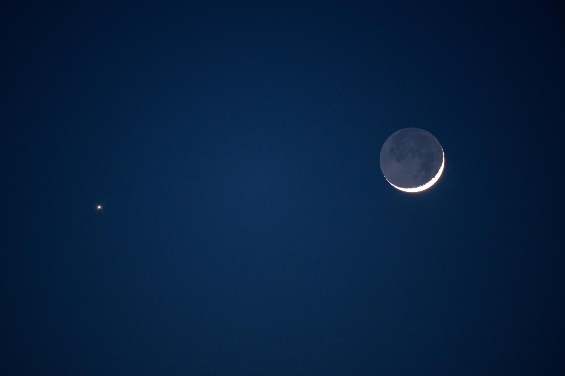 Una Luna en fase muy fina sobre un cielo crepuscular azul oscuro: solo brilla un arco de luz en su borde inferior derecho, y el resto del disco se adivina en gris gracias a la luz cenicienta. A su izquierda y algo más abajo, Venus aparece como un punto blanco intenso y aislado.