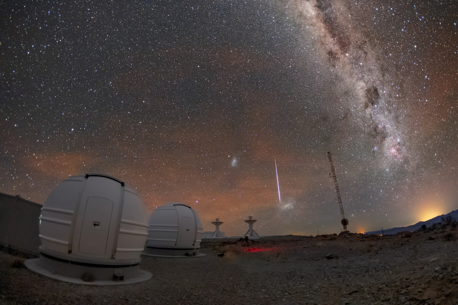 Una raya fina de luz blanca baja casi vertical por un cielo lleno de estrellas, con la banda de la Vía Láctea cruzando arriba a la derecha, por encima de dos cúpulas blancas y pequeñas de un observatorio levantado en un desierto de piedra.