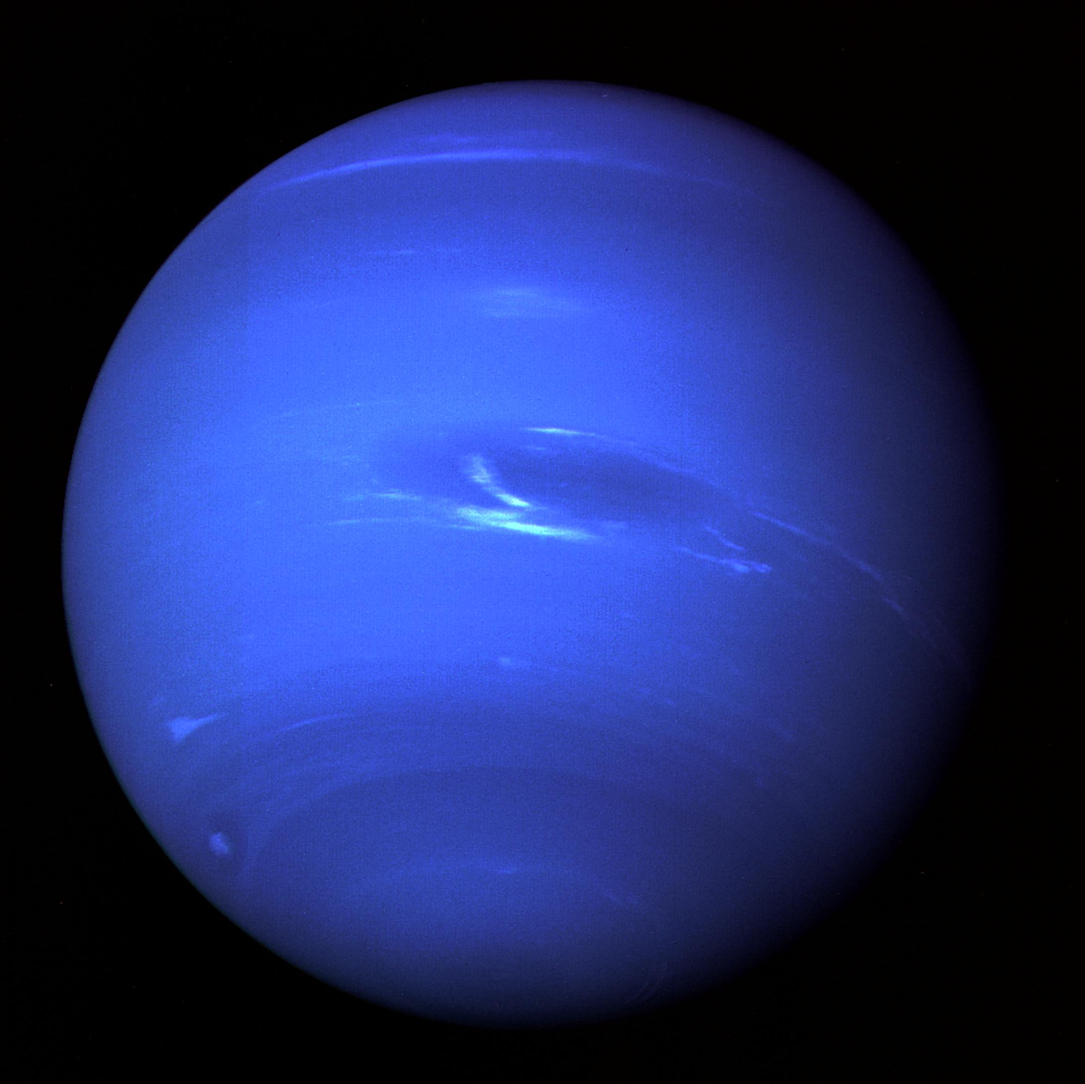 El disco de Neptuno fotografiado por la sonda Voyager 2: una esfera azul intensa cruzada por bandas horizontales de distinto tono, con una gran mancha oscura ovalada acompañada de nubes blancas alargadas y un pequeño penacho brillante cerca del borde.