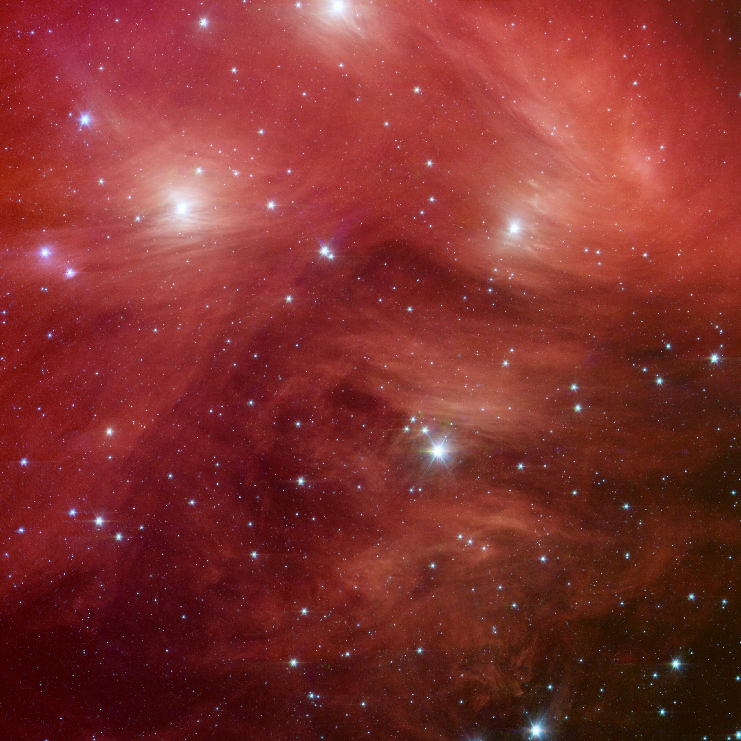 Imagen infrarroja del cúmulo de las Pléyades tomada por el telescopio espacial Spitzer: sobre un fondo rojo intenso surcado por velos y filamentos de polvo se reparten cientos de estrellas como puntos blancos, y media docena de ellas, mucho más brillantes, muestran destellos de difracción en forma de cruz. Los colores no son los que vería el ojo: la NASA los asignó a distintas longitudes de onda del infrarrojo.