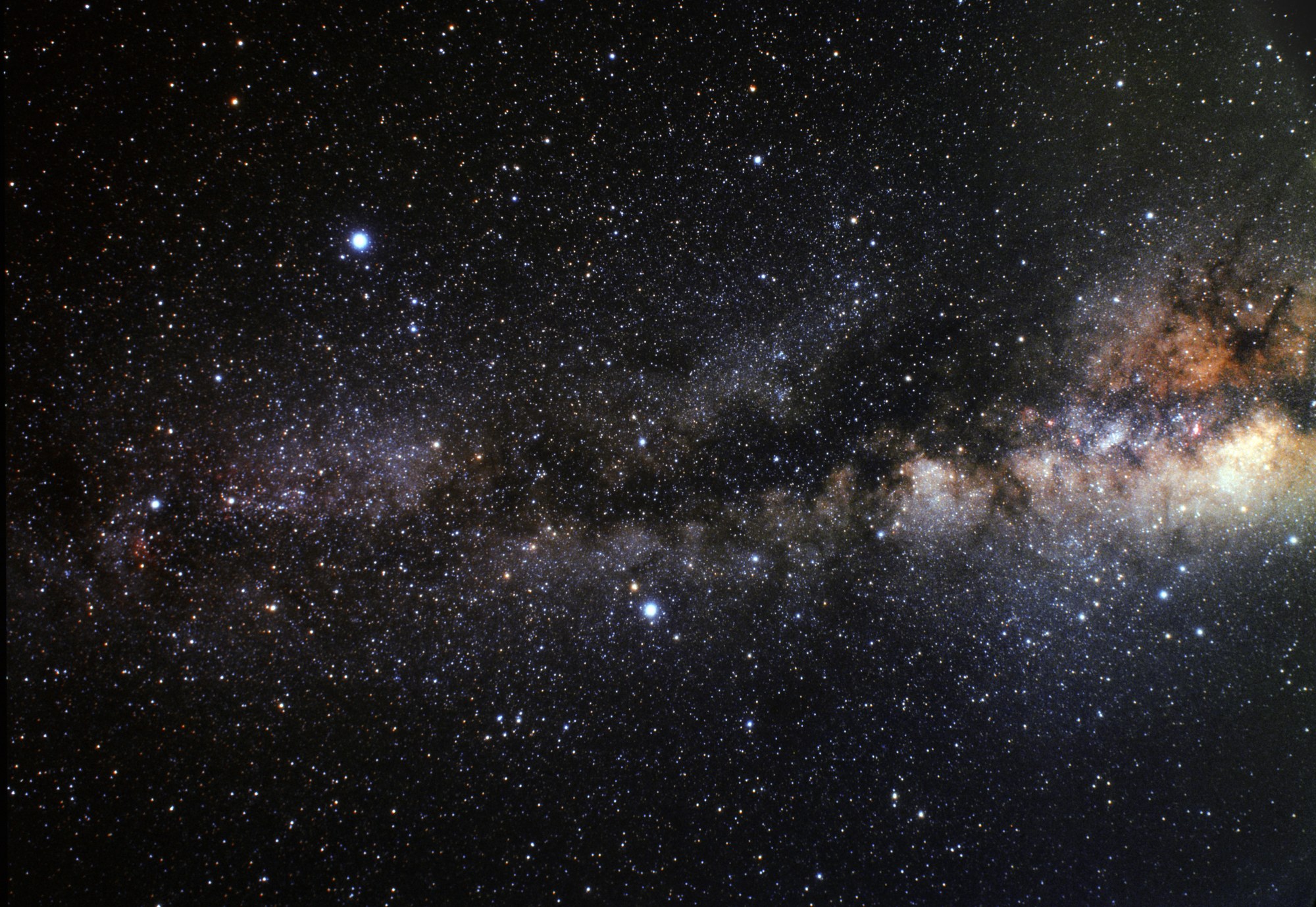 Fotografía de campo muy amplio del cielo nocturno, con miles de estrellas sobre fondo negro. La banda de la Vía Láctea la cruza en diagonal, más ancha y dorada hacia la derecha, donde se apelotonan nubes de estrellas, y más tenue hacia la izquierda, partida a lo largo por vetas oscuras de polvo. Tres estrellas destacan sobre las demás por su tamaño y su brillo blanco azulado: una arriba a la izquierda, otra abajo en el centro y una tercera en el borde izquierdo, metida en la banda luminosa.