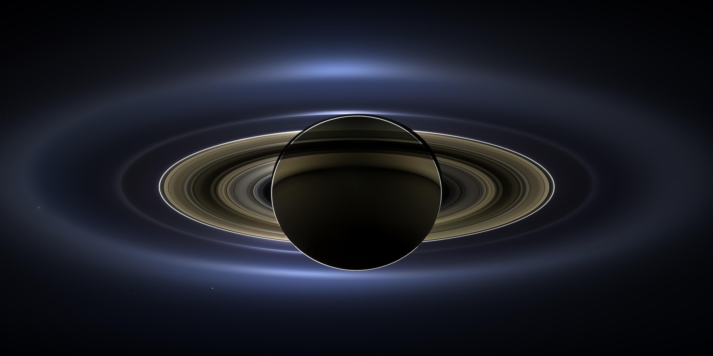 Saturno a contraluz con el Sol escondido detrás: el disco del planeta en sombra, rodeado por los anillos iluminados en tonos dorados y por un halo azulado de anillos exteriores sobre el fondo negro.
